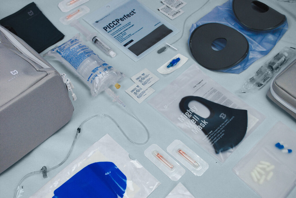 Many Mighty Well products laying flat on a blue background - Fluid Motion Backpack, PICCPerfect PICC Line Cover, Mighty Well Mask, H2O Shield and various medical supplies. 